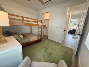 A compact bedroom with a bunk bed, a bedside table with a lamp, a green carpet, and a hallway leading to a bathroom. - Grown Up Queen Bunk Cottage | Sleeps 4-6 | Pool + River (New Braunfels)