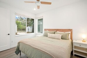 A bedroom with a bed, a nightstand, a lamp, a ceiling fan, and a window with a view of a house and trees. - River Cottage Sleeps 2-4 | Pool + Pickleball (New Braunfels)
