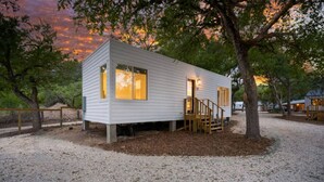 A single-story cabin with a wooden staircase, large windows, and a gravel ground. - Hill Country Cottage | 1BR King | Pool + Hot Tub | Pets Welcome (New Braunfels)