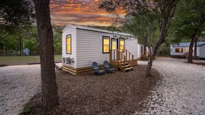 A small, single-story mobile home with a porch and a staircase, surrounded by trees and gravel. - Guadalupe Tiny Cabin | Pool+Hot Tub | NBTX (New Braunfels)