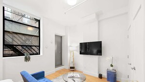 Uma sala de estar moderna com um sofá azul, uma mesa de centro de vidro, uma TV na parede e uma janela com vista para um edifício de tijolos.