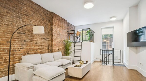 A modern living room with a brick wall, a white sofa, a coffee table, and a staircase leading to an upper level. - Charming UES 1BR Duplex Near Central Park (New York)