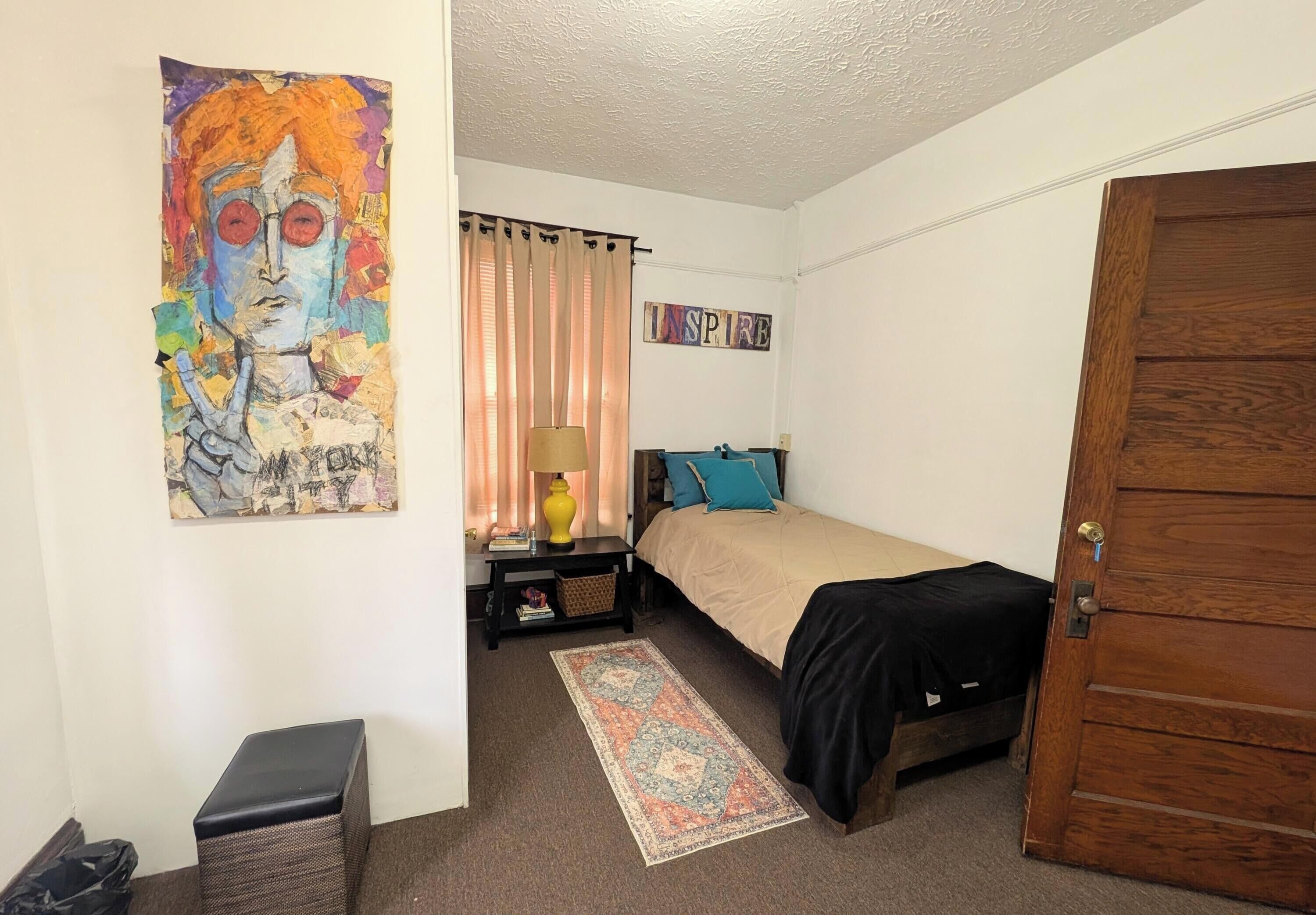 A bedroom with a bed, a nightstand, a lamp, a rug, a wooden door, and a painting on the wall.