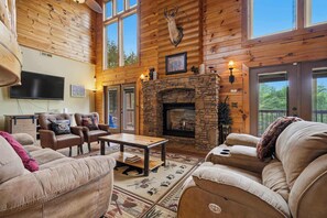 A log cabin interior with a stone fireplace, leather furniture, and large windows. - 4BR Cabin w/ Yard, Office, Hot Tub & Firepit! (Sevierville)