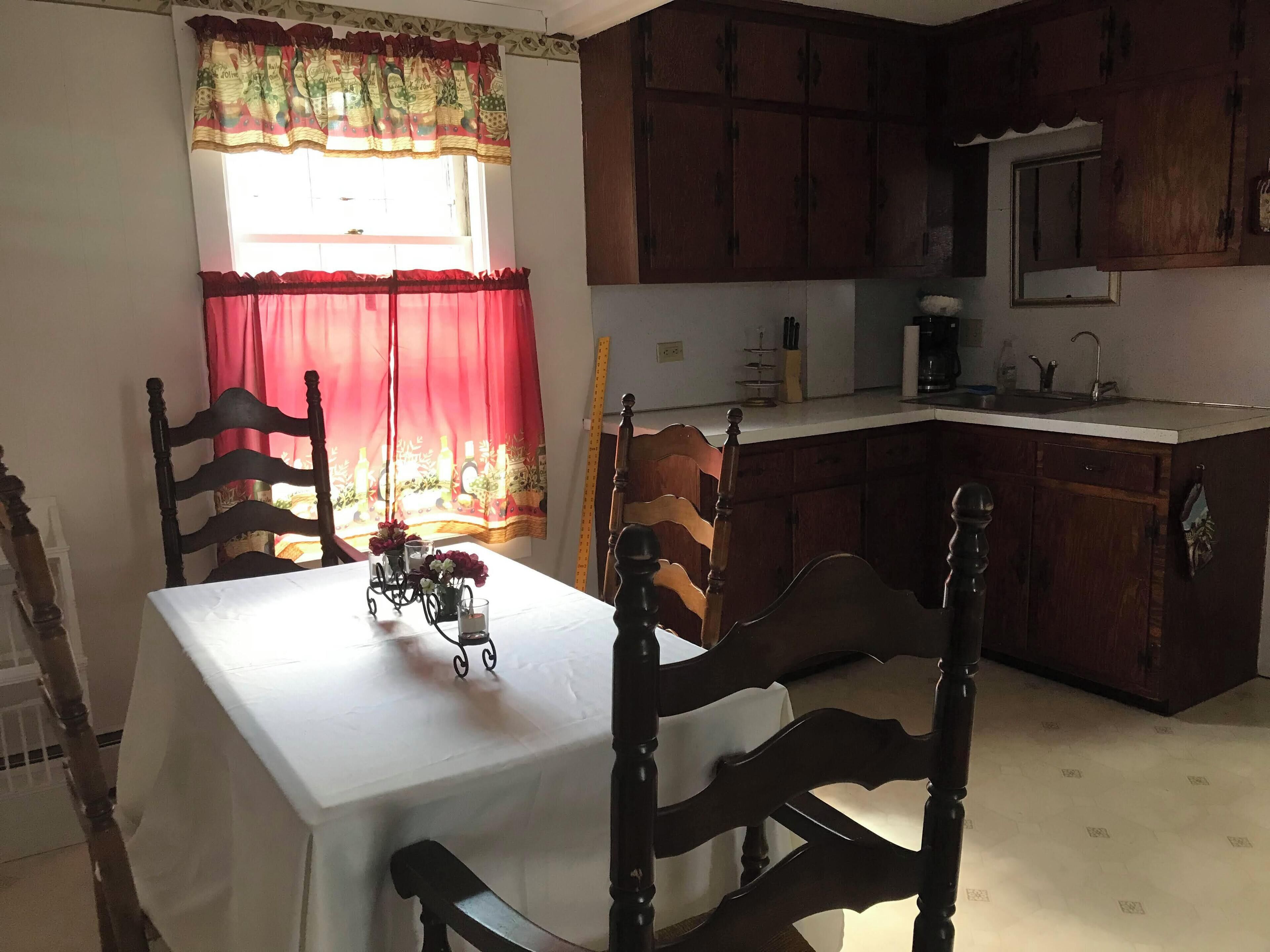 Un comedor con una mesa preparada para dos personas, sillas de madera, una ventana con cortinas rojas y una cocina al fondo.