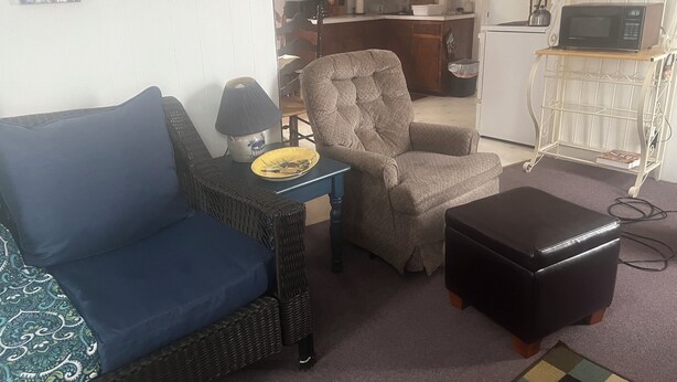 Een woonkamer met een bank, fauteuil en voetenbankje, een tafeltje met een lamp, en een keuken in de achtergrond.