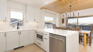 A modern kitchen with white cabinets, a dishwasher, and a large island. There is a dining area with a table and chairs, and a view of mountains through large windows. - *New to Market* 5 Bedroom Big Sky Resort Condo (Big Sky)