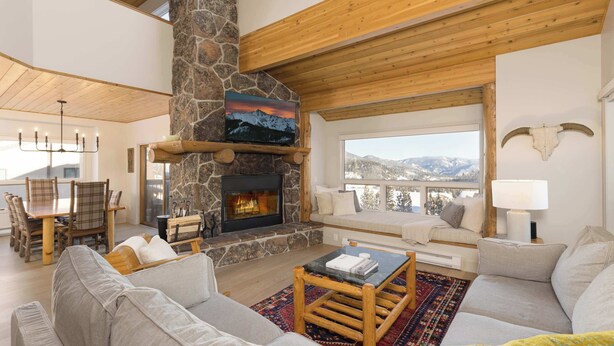 A cozy living room with a stone fireplace, wooden beams, and a large window overlooking a snowy mountain landscape. - *New to Market* 5 Bedroom Big Sky Resort Condo (Big Sky)