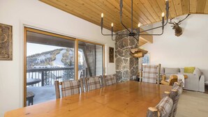 A spacious dining area with a large wooden table and plaid chairs, a stone accent wall, and a view of a snowy mountain landscape through large sliding doors. - *New to Market* 5 Bedroom Big Sky Resort Condo (Big Sky)
