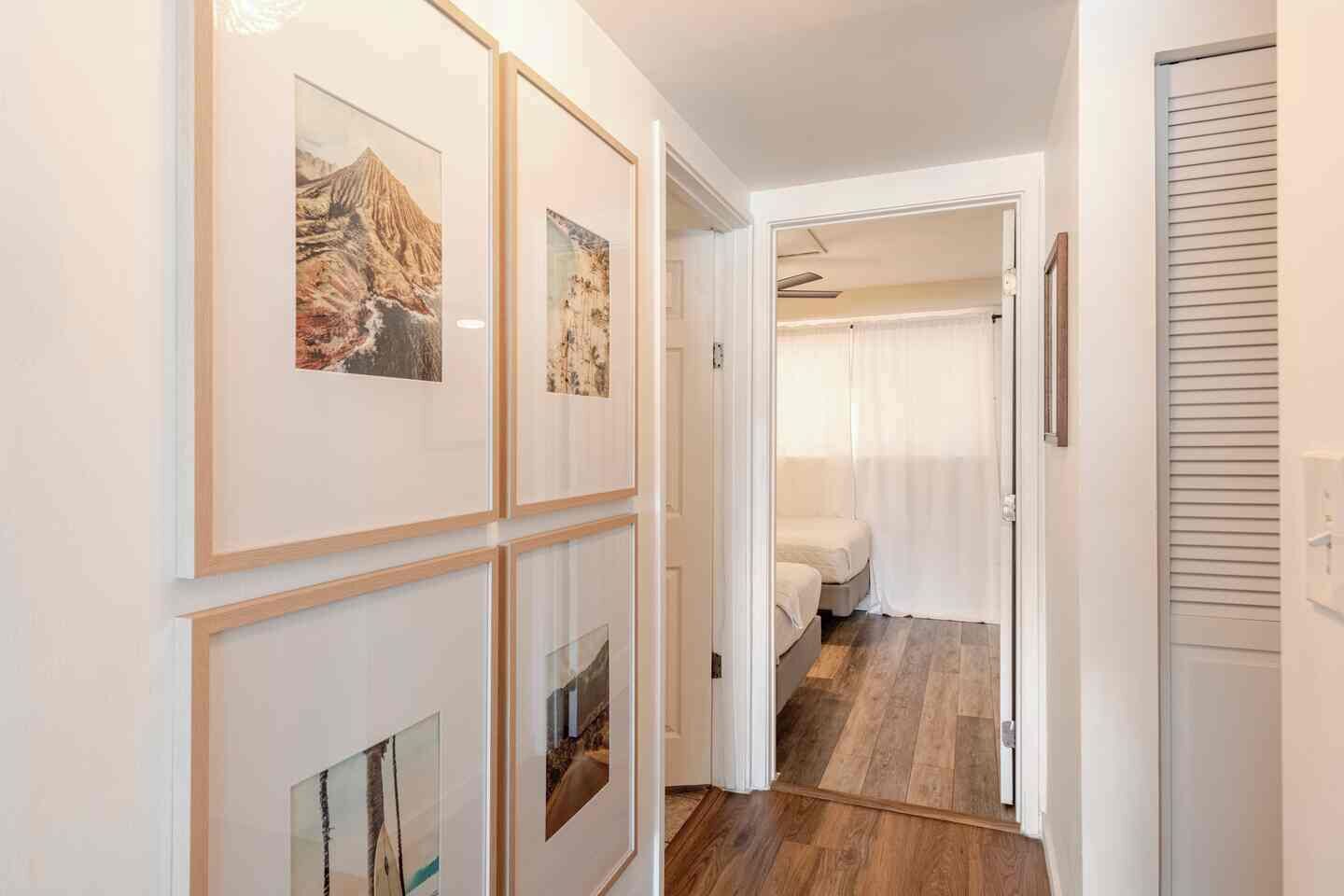 A hallway with framed pictures on the wall, a bed in the adjacent room, and a wooden floor.