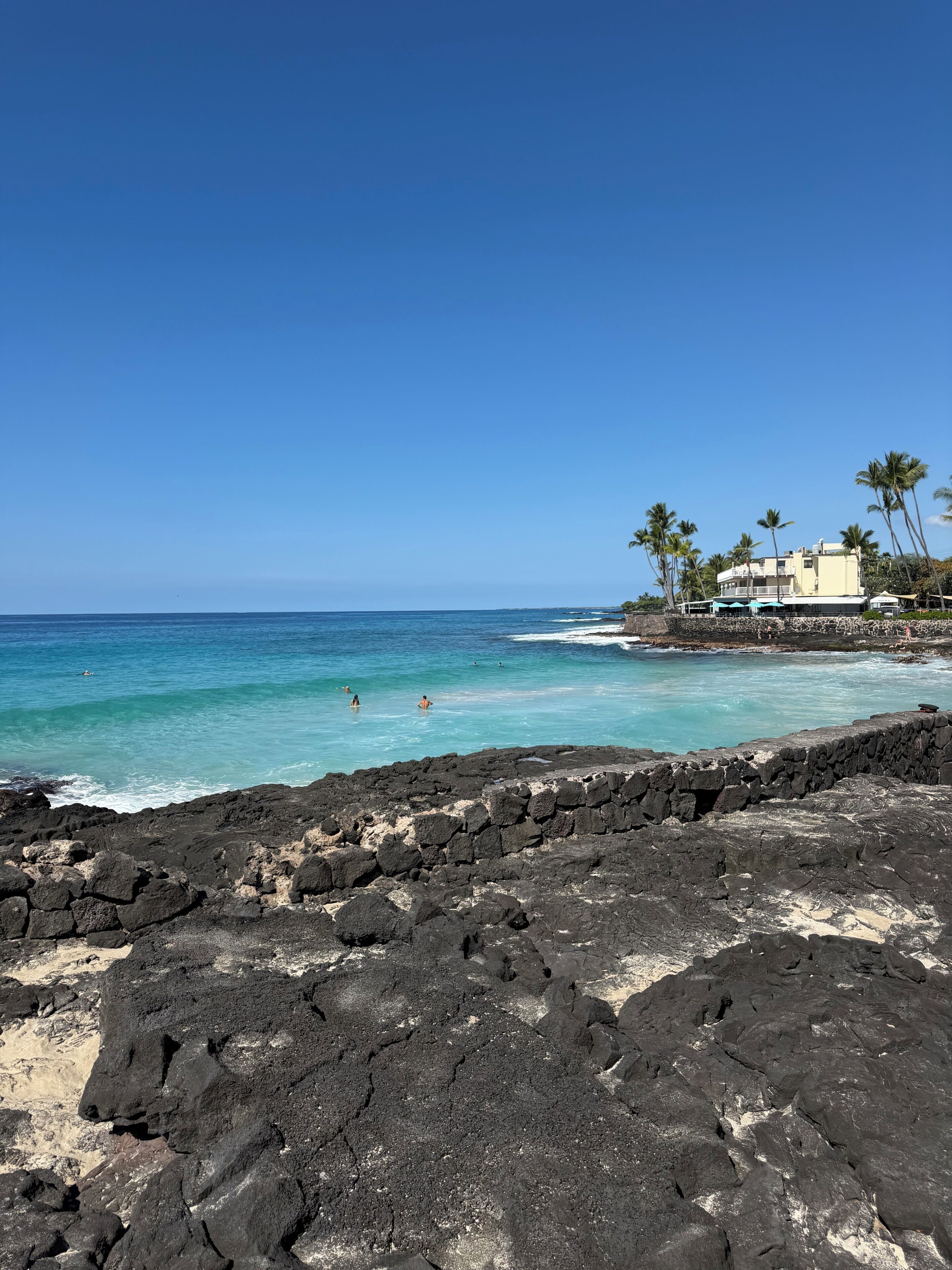 Une plage avec une eau d’un bleu limpide, un premier plan rocheux et un bâtiment doté d’un balcon.
