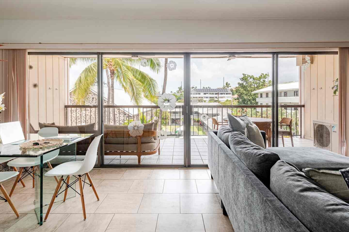 A modern living room with a glass door leading to a balcony, a sofa, a dining table with chairs, and a view of palm trees and buildings.