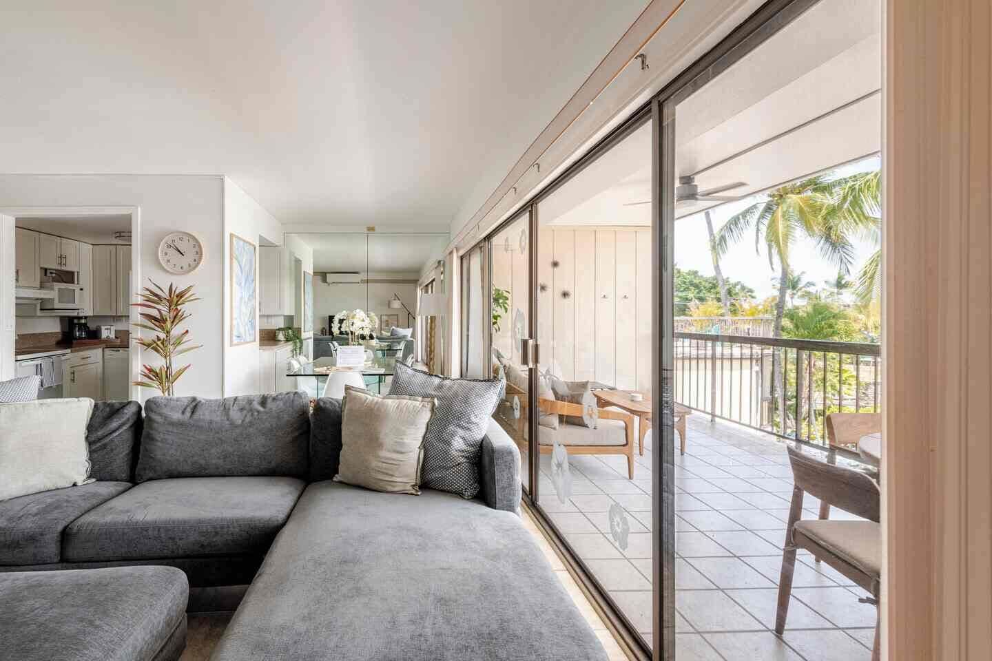 Un salon moderne avec un canapé d’angle gris, un coin repas doté d’une table en verre et un balcon aménagé avec du mobilier d’extérieur.