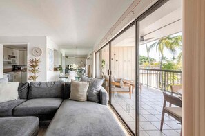 A modern living room with a grey sectional sofa, a dining area with a glass table, and a balcony with outdoor furniture. - Peek-a-Boo Ocean View Condo in Kona - Walk to Beaches, Shops & Restaurants (Kailua Kona)