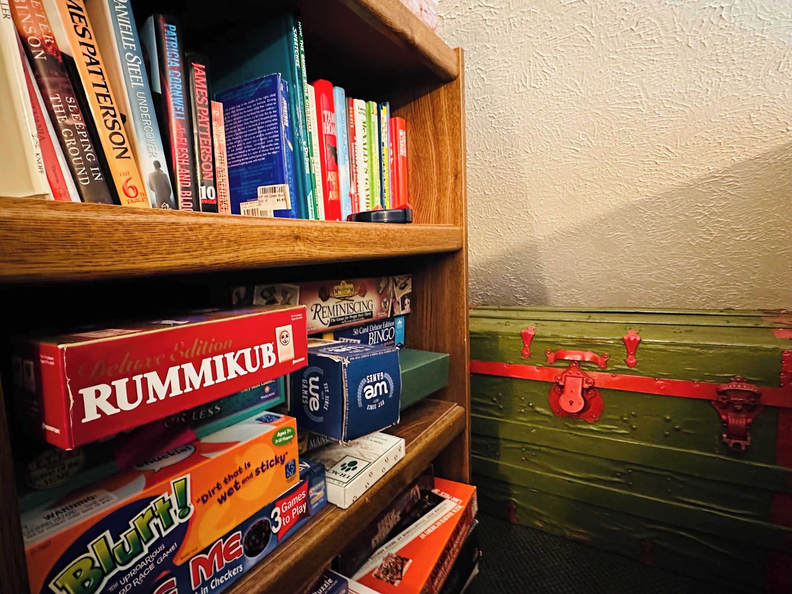 A bookshelf with books and games, including a red box labeled "RUMMIKUB" and a green trunk with red straps.