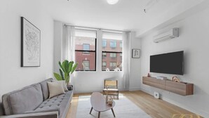Uma sala de estar moderna com um sofá cinzento, uma mesinha redonda pequena, uma janela grande com cortinas, uma televisão de tela plana e uma estante com objetos decorativos.