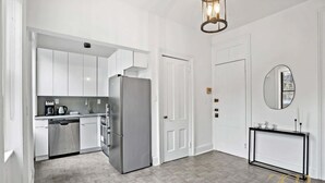 A modern kitchen with white cabinets, a stainless steel refrigerator, and a dishwasher. There is a hallway with a door leading to another room. - Sun-Filled Brownstone Apt in Prime Chelsea (New York)