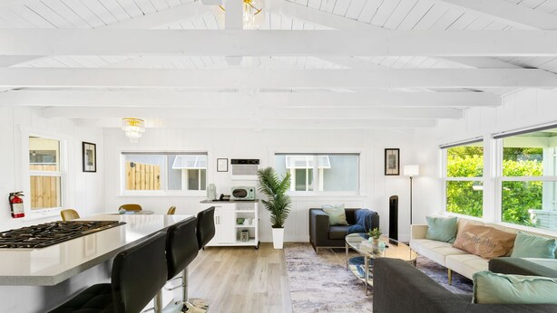 Eine moderne Küche mit einer zentralen Kochinsel, ein Essbereich mit Barhocker, ein Wohnzimmer mit Sofa und Couchtisch sowie große Fenster, die für natürliches Licht sorgen.