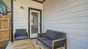 Een overdekt terras met een houten tafel, twee rieten stoelen met kussens, en een deur met een raam.