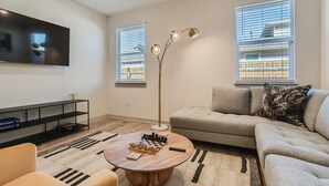 Een woonkamer met een flatscreen televisie, een vloerlamp, een bank, een bijzettafeltje met een schaakspel en een boekenkast.