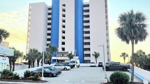 Ein mehrstöckiges Hotel mit einer markanten blau-weißen Fassade, einem Parkplatz mit mehreren Autos und Palmen im Vordergrund.