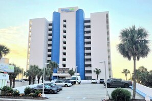 Ein mehrstöckiges Hotel mit einer markanten blau-weißen Fassade, einem Parkplatz mit mehreren Autos und Palmen im Vordergrund.
