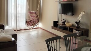 A spacious living room with a ceiling fan, a hanging chair, a TV on a stand, a dining table with chairs, and a large window with curtains. - Your best spot in Ipanema, just steps from the sea and the lagoon. (Rio de Janeiro)