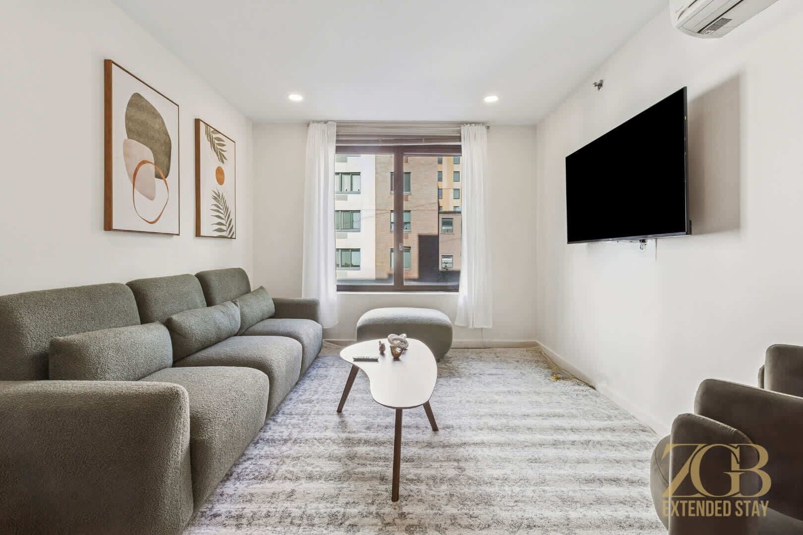 Un salon moderne avec un canapé gris, une petite table blanche et un téléviseur à écran plat fixé au mur.