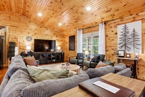A log cabin interior with a sofa, chairs, a TV, and a window with a view. - Newly Refreshed : Knotty Pine Cabin + Hot Tub (Logan)