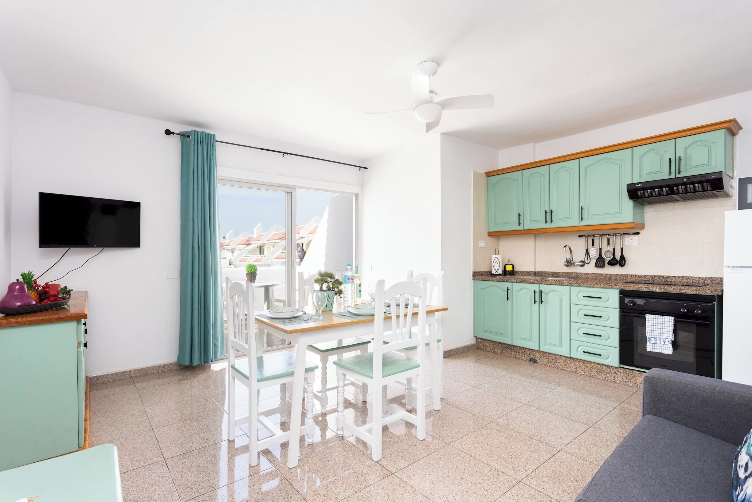 A modern kitchen with teal cabinets, a dining area with white chairs, and a flat-screen TV mounted on the wall. - Home2Book Relax & Enjoy in Las Americas (Playa de la Américas)