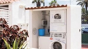 A compact laundry facility with a washing machine, a drying machine, and a blue container, surrounded by a white wall and a small garden with aloe plants. - Home2Book Relax & Enjoy in Las Americas (Playa de la Américas)
