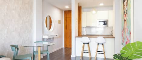 A modern kitchen with white cabinets, a microwave, and a wooden door. There is a round table with chairs and a large plant in the foreground.