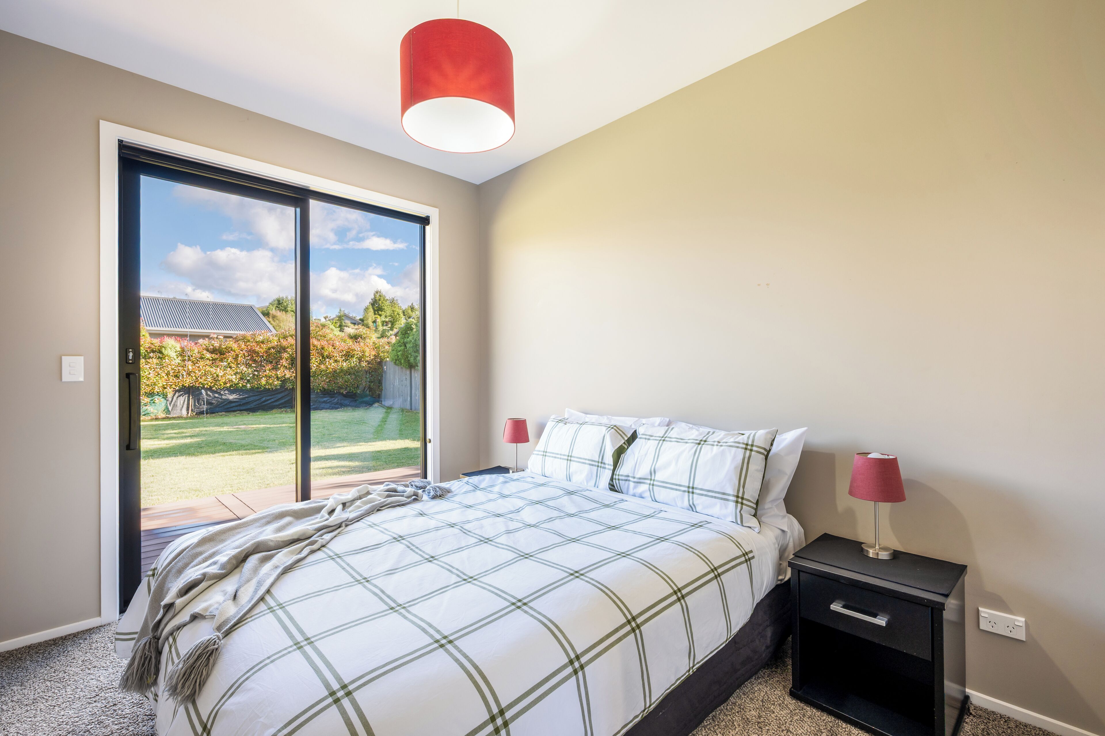 A bedroom with a large bed, two bedside tables, a red pendant light, and a sliding glass door leading to an outdoor area.