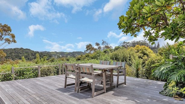 Une terrasse en bois avec une table et des chaises, entourée d’une végétation luxuriante et avec des collines en arrière-plan.