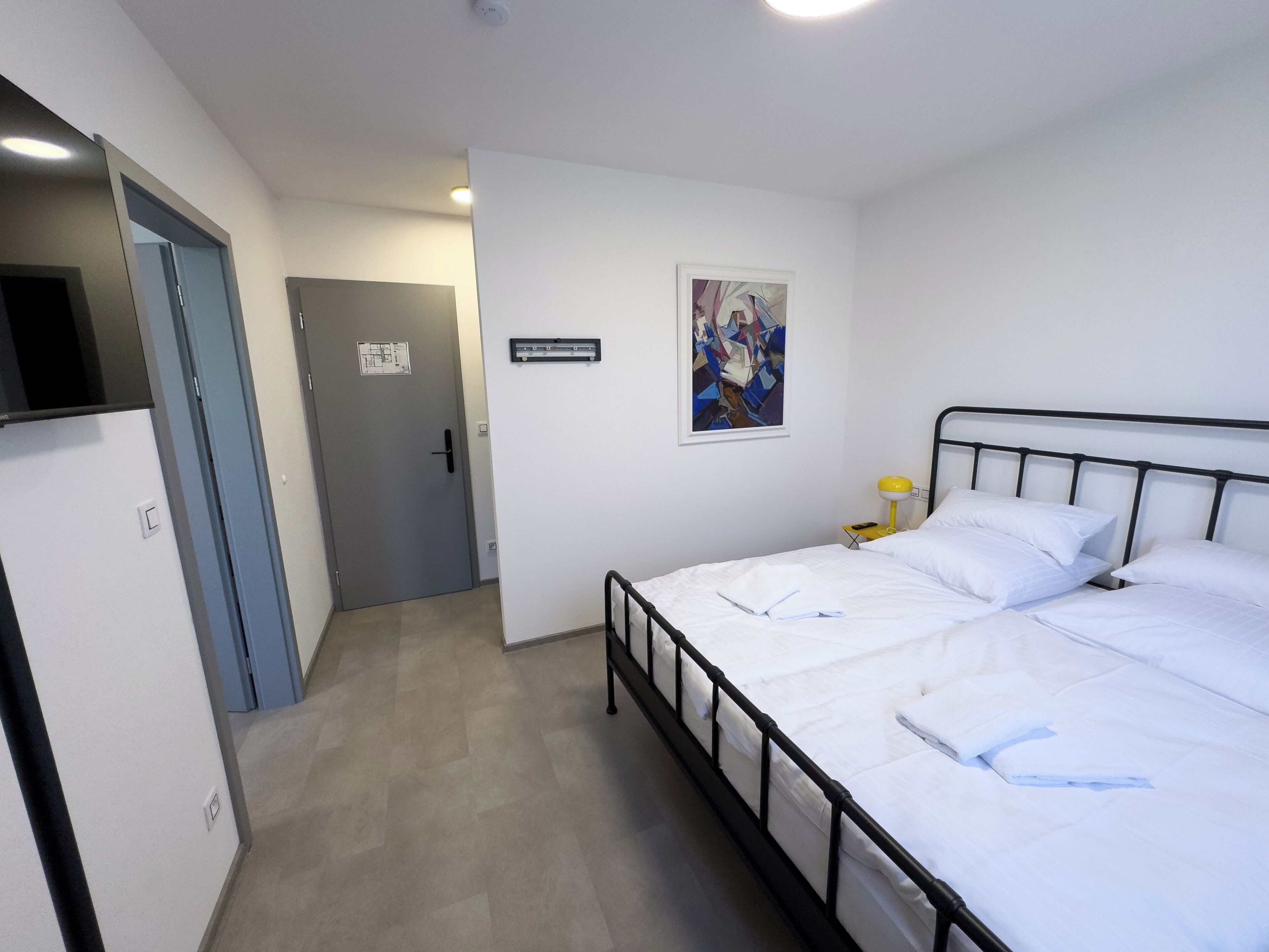 Un dormitorio con una cama, una mesita de noche con lámpara, una puerta y una obra de arte colgada en la pared.