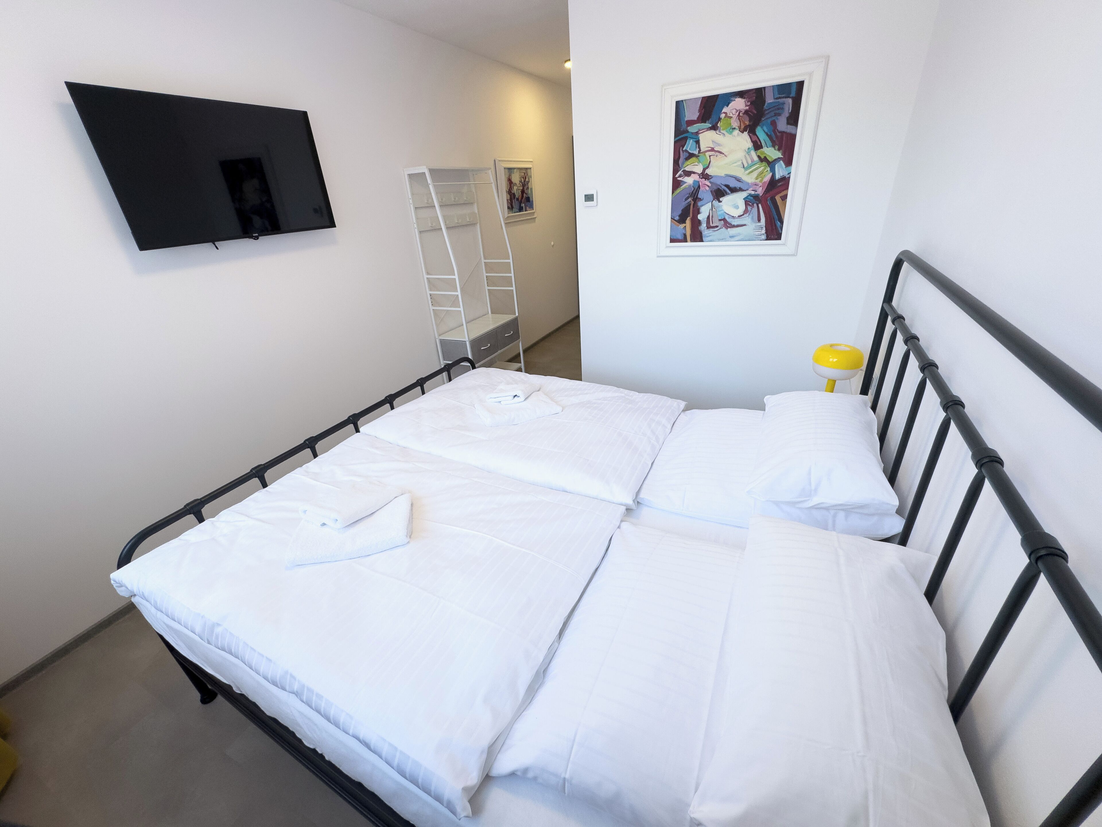 Una cama bien hecha con sábanas blancas, un cabecero de metal negro, una mesita de noche con una lámpara amarilla, un televisor blanco montado en la pared y una escalera blanca apoyada contra la pared.