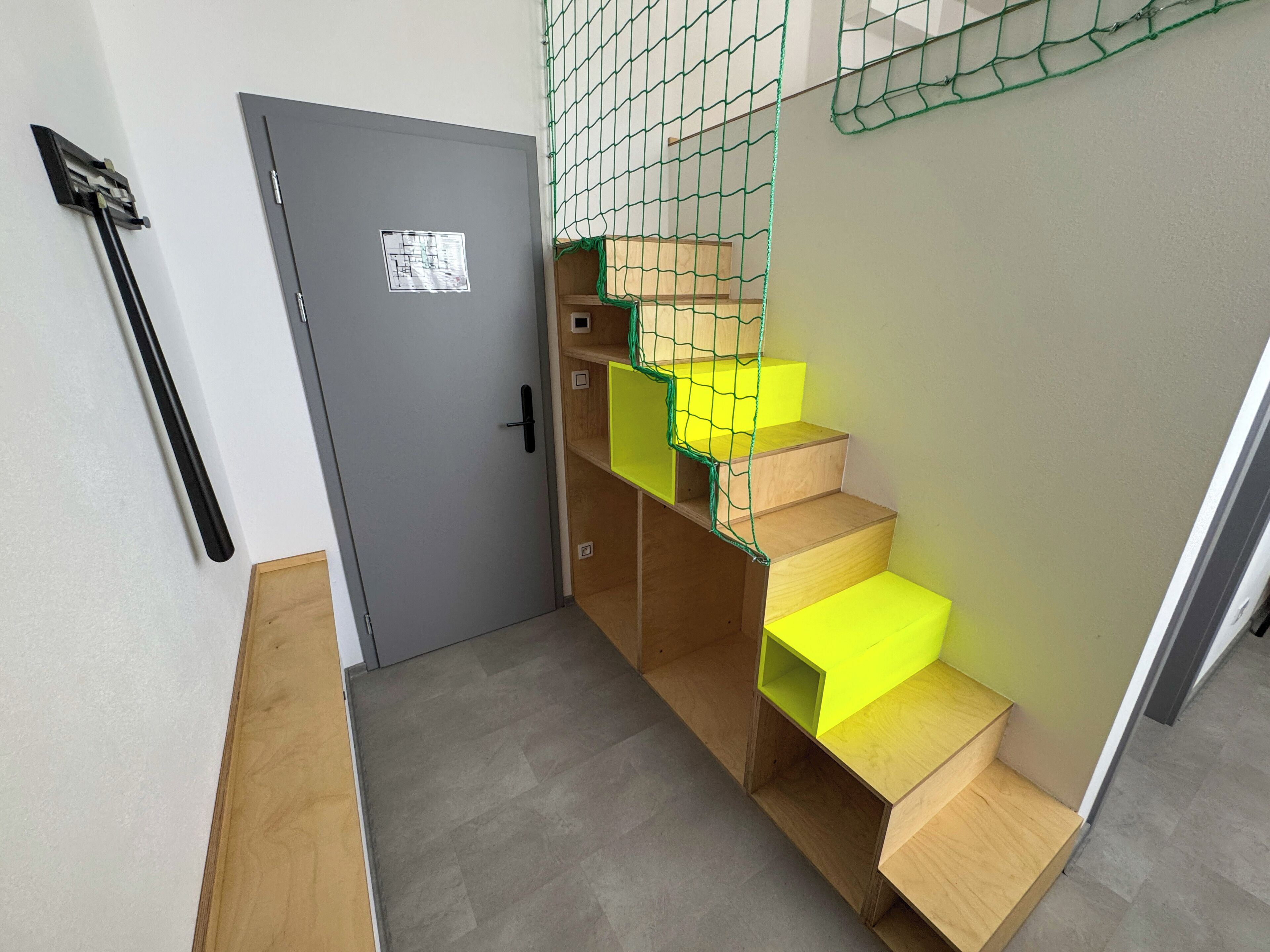 Eine Treppe mit Holzsitzstufen und leuchtend gelben Trittstufen, einer grauen Tür und einer grünen Netzanlage.