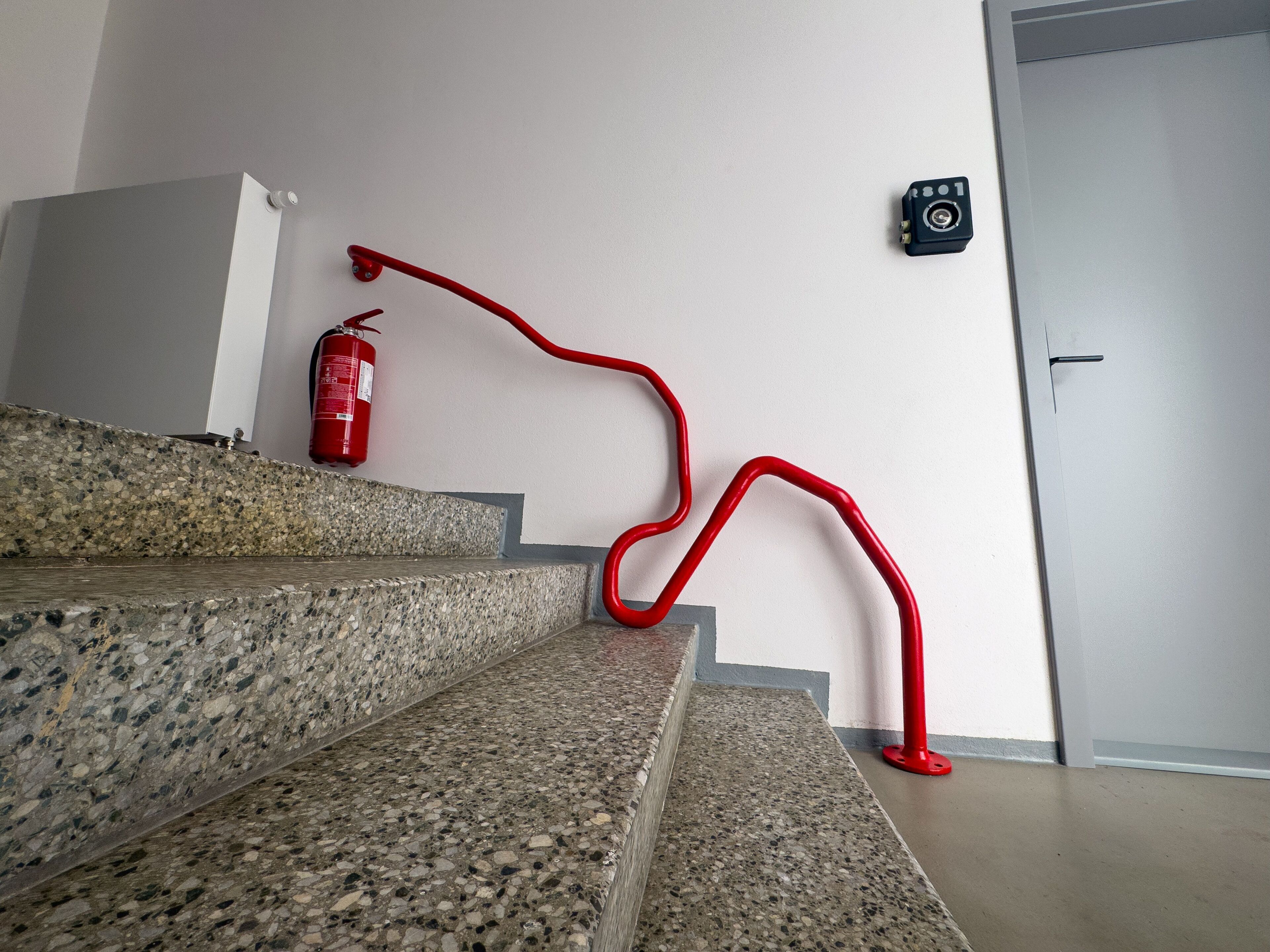A staircase with granite steps and a red handrail, a fire extinguisher mounted on the wall, and a door with a security camera.