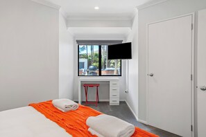 Una habitación de hotel moderna con una cama, un edredón naranja, un escritorio con una silla, un televisor montado en la pared y una ventana con vistas a palmeras.