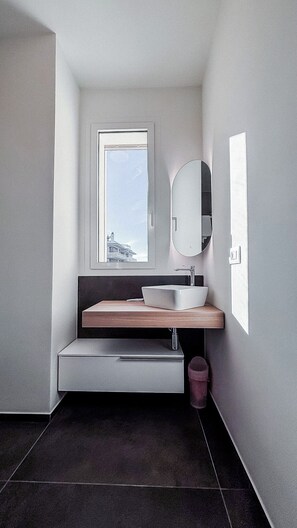 A modern bathroom with a large window, a rectangular sink, a round mirror, and a dark floor.