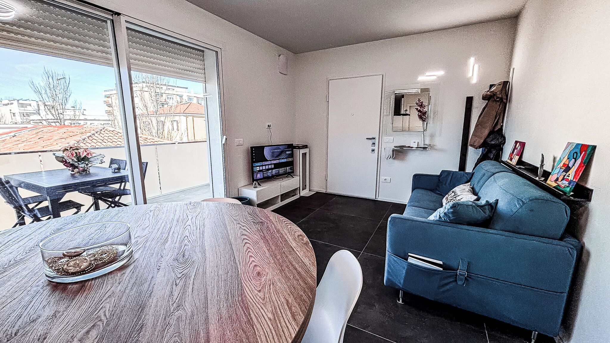 A modern living room with a round wooden table, a blue sofa, a TV, and a balcony with a view of buildings.