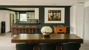 Une cuisine moderne avec un mur de couleur sombre, une grande table à manger en bois et des chaises noires. On peut voir un vase avec des fleurs sur la table et un tableau au-dessus.