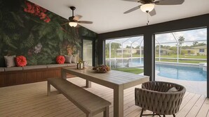 Un salon moderne avec un plancher en bois, une table à manger, un banc, un canapé avec des coussins, un ventilateur de plafond et une grande fenêtre donnant sur une piscine.