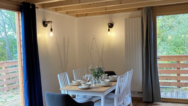 Un coin repas avec une table en bois dressée pour quatre personnes, entourée de chaises blanches. On y trouve deux appliques murales, une fenêtre donnant sur un paysage verdoyant et une grande porte en bois.