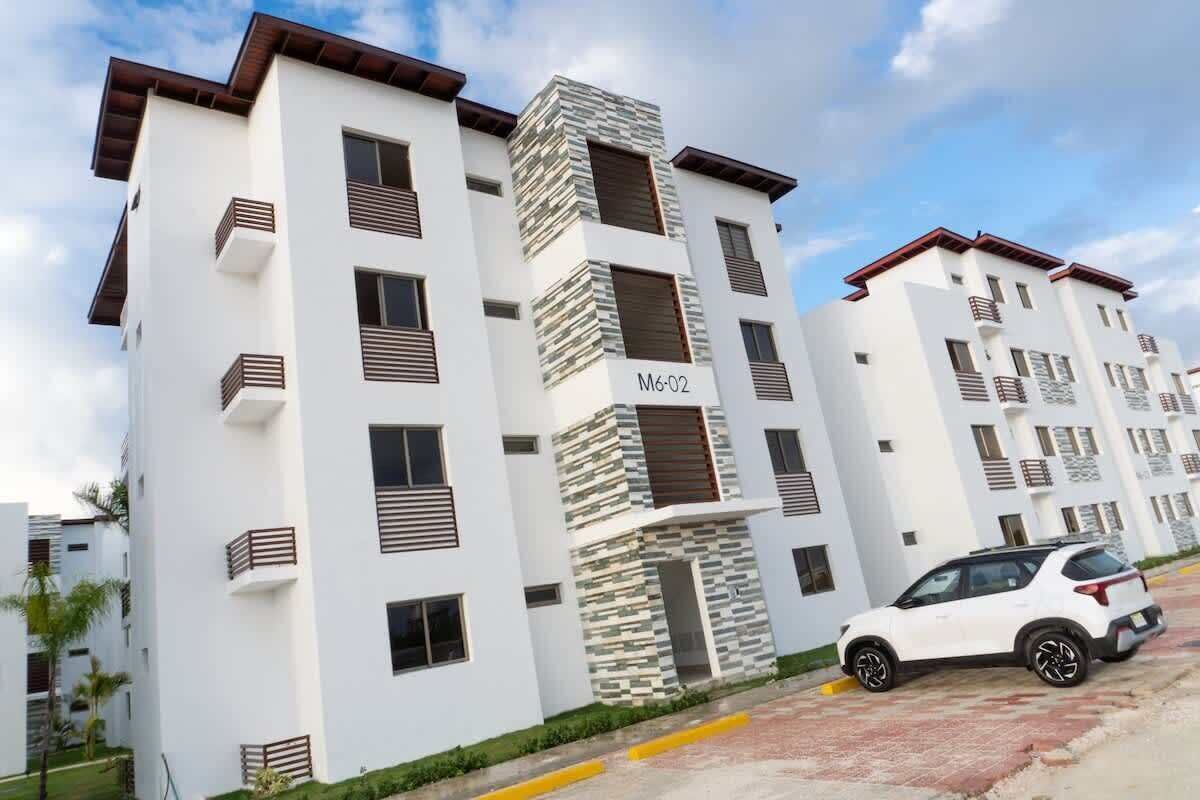 A modern multi-story residential building with balconies, a car parked in front, and a clear sky.