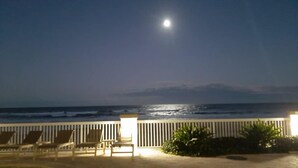 A beach view with a moonlit sky, a white fence, and lounge chairs. - Beachfront studio w/pool & fire pit (Daytona Beach)