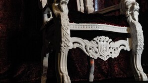 A white ornate chair with intricate carvings and a lion head design, positioned against a dark red curtain backdrop. - Hourly hotel BlackKingdom (Freiburg im Breisgau)