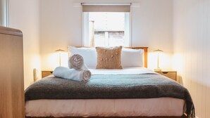 A neatly made bed with a rolled towel, a decorative pillow, and a blanket. There are two bedside lamps and a window with a view of buildings. - The Chance - Immerse yourself in the oceanic ambience of South Beach. (Port Fairy)