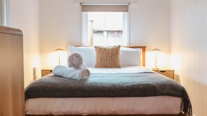 Ein ordentlich bezogenes Bett mit einem zusammengerollten Handtuch, einem Zierkissen und einer Decke. Zwei Nachttischlampen und ein Fenster mit Blick auf Gebäude sind vorhanden.