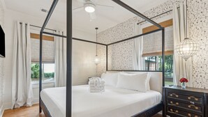 Un lit à baldaquin avec du linge de lit blanc, une table de chevet en bois foncé dotée de tiroirs et un ventilateur de plafond.